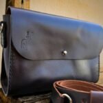 Close do fechamento frontal da bolsa Verona Caribu com detalhe da logomarca timbrada no couro café.